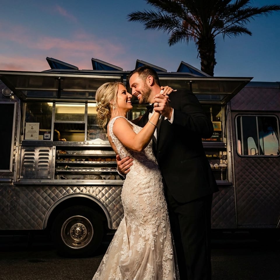 A happy couple dances in front of a food truck at sunset, palm trees in the background.