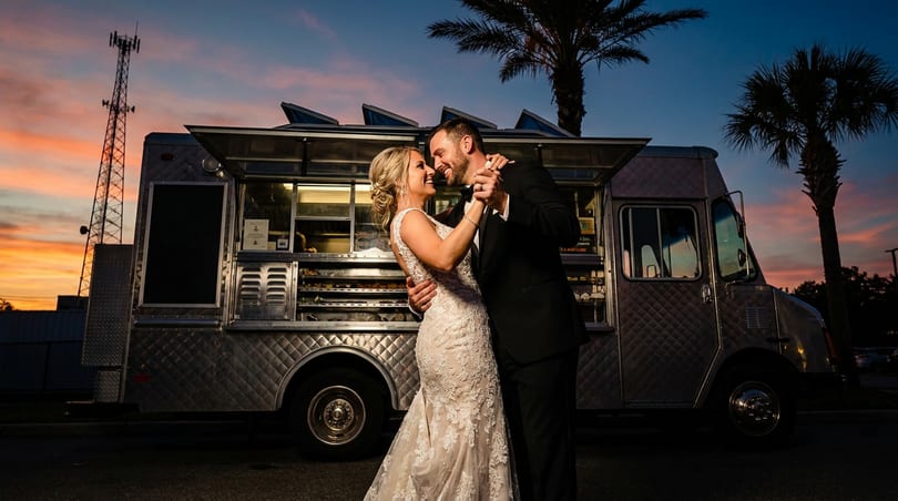 A happy couple dances in front of a food truck at sunset, palm trees in the background.