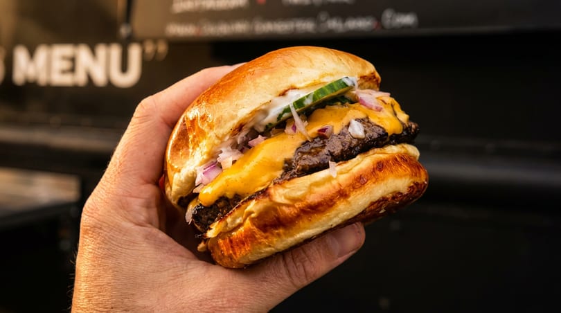 Hand holding a cheeseburger with melted cheese, red onions, and cucumber on a brioche bun.