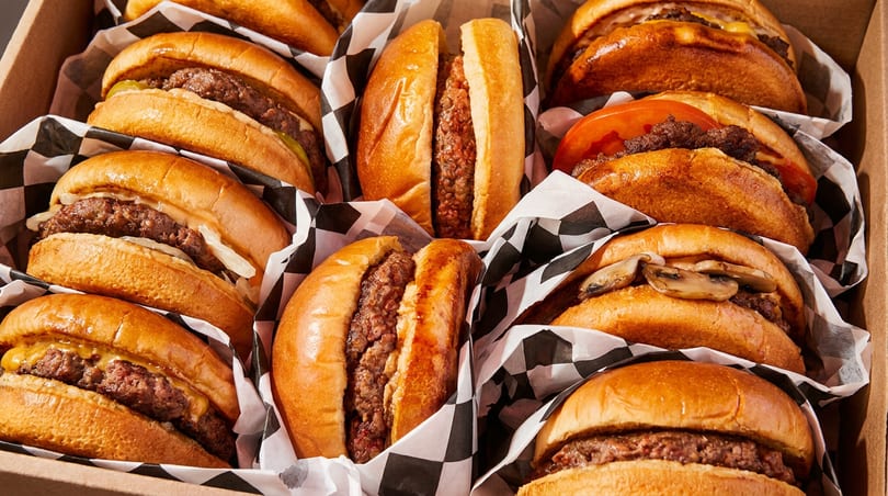 A box filled with many small sliders, some with cheese, tomato, or mushrooms, wrapped in checkered paper.