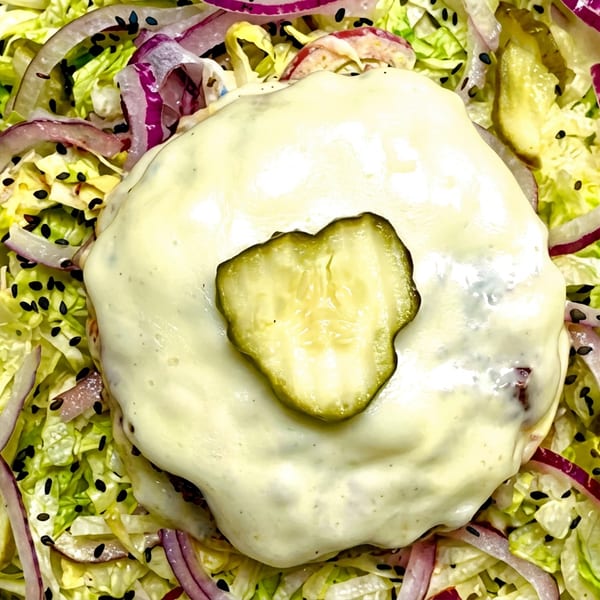 Cheeseburger patty with a pickle slice on top, surrounded by lettuce, red onion, and sesame seeds.