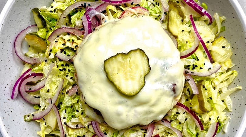 Cheeseburger patty with a pickle slice on top, surrounded by lettuce, red onion, and sesame seeds.