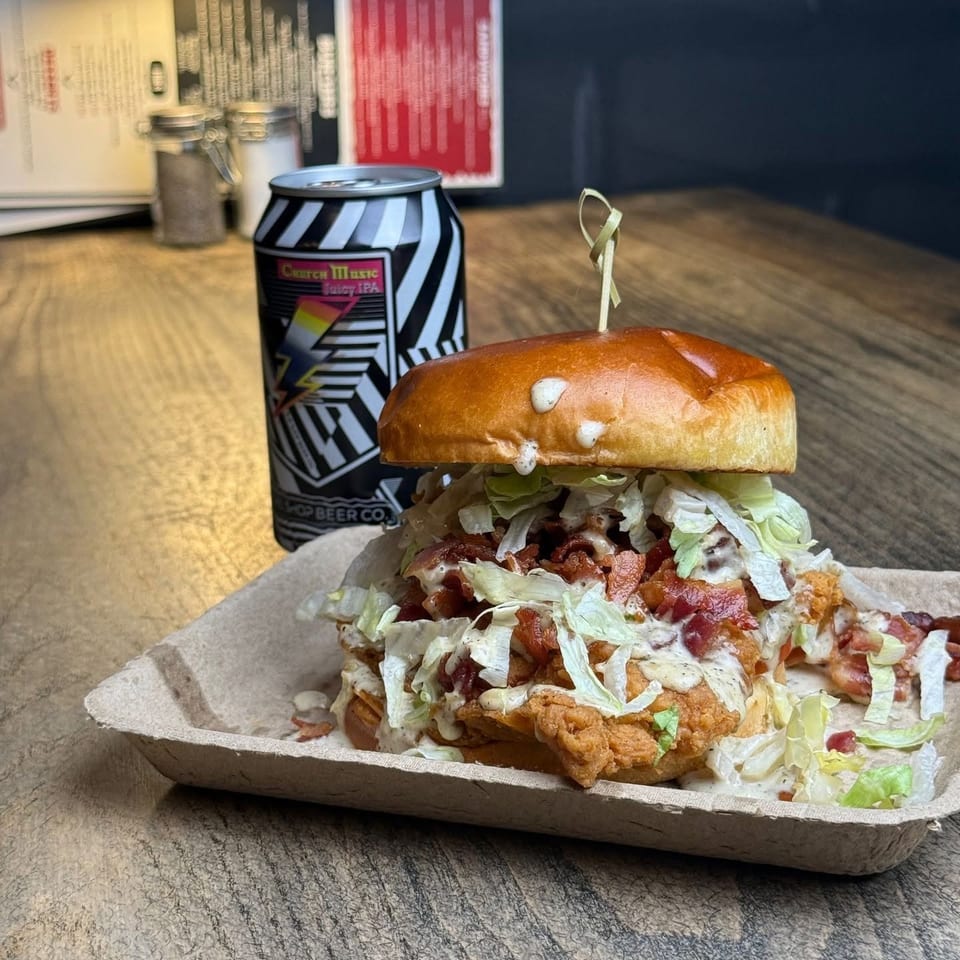 Crispy chicken sandwich with bacon, lettuce, and sauce on a paper tray, next to a Church Music IPA.