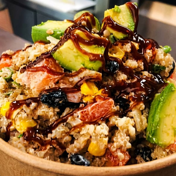A colorful quinoa bowl with avocado, corn, berries, and red fruit, drizzled with sauce.