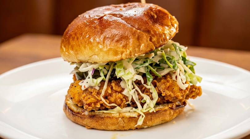 Crispy fried chicken sandwich with coleslaw and pickles on a brioche bun, served on a white plate.